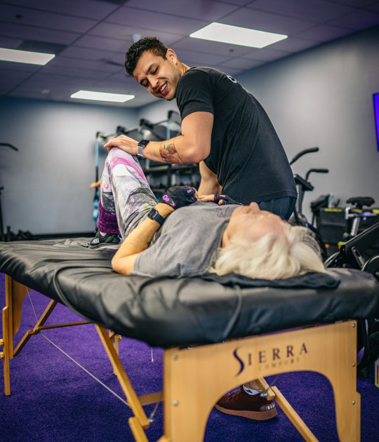 Assisted stretching session at Ageless Fitness Encinitas, with a coach guiding a senior woman through a gentle leg stretch on a treatment table in a clean, modern gym.