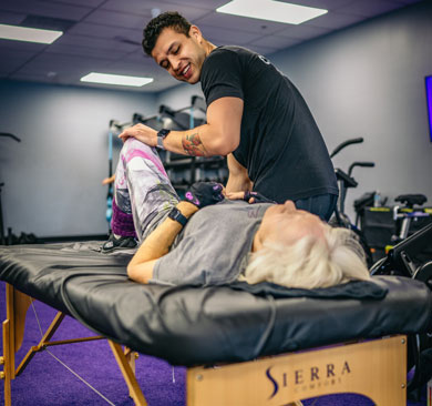 Coach assisting an older adult with guided stretching on a treatment table to improve mobility and joint flexibility in a gym setting.